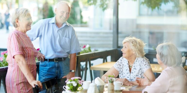 Group of Seniors Socializing at an Outdoor Café Table of an Assisted Living Community, All Smiling and Engaged in Cheerful Conversation